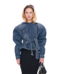 A model wearing a blue denim jacket with a unique draped, oversized silhouette, a self-tie belt at the waist, and flap chest pockets, paired with a black skirt and holding a black handbag against a white background.