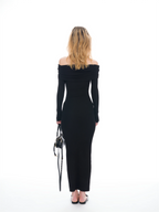 A woman viewed from behind wearing a fitted off-the-shoulder long-sleeve maxi dress in solid black, holding a small black handbag against a plain white studio background.