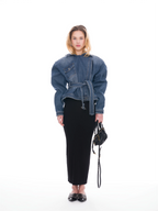 A model stands against a white studio background wearing a blue denim belted jacket with voluminous sleeves, a long black skirt, black pointed flats, and holding a small black handbag.