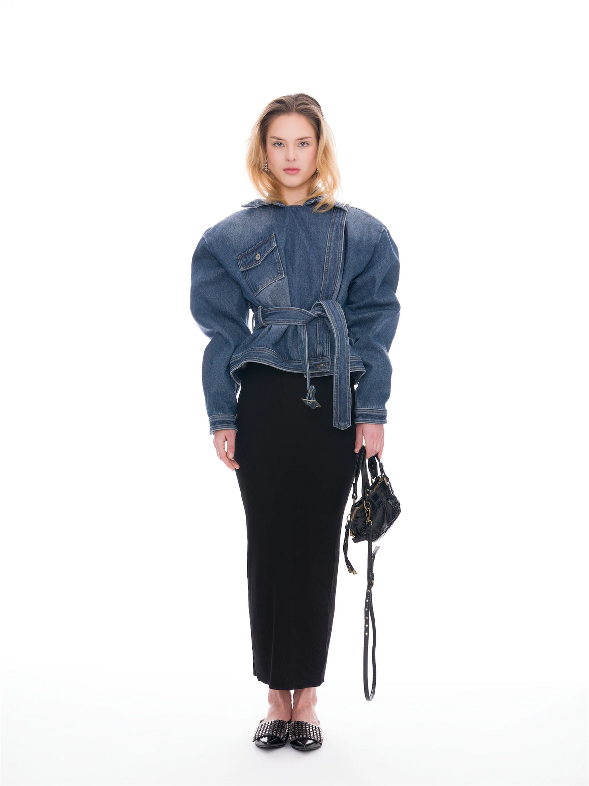 A model stands against a white studio background wearing a blue denim belted jacket with voluminous sleeves, a long black skirt, black pointed flats, and holding a small black handbag.