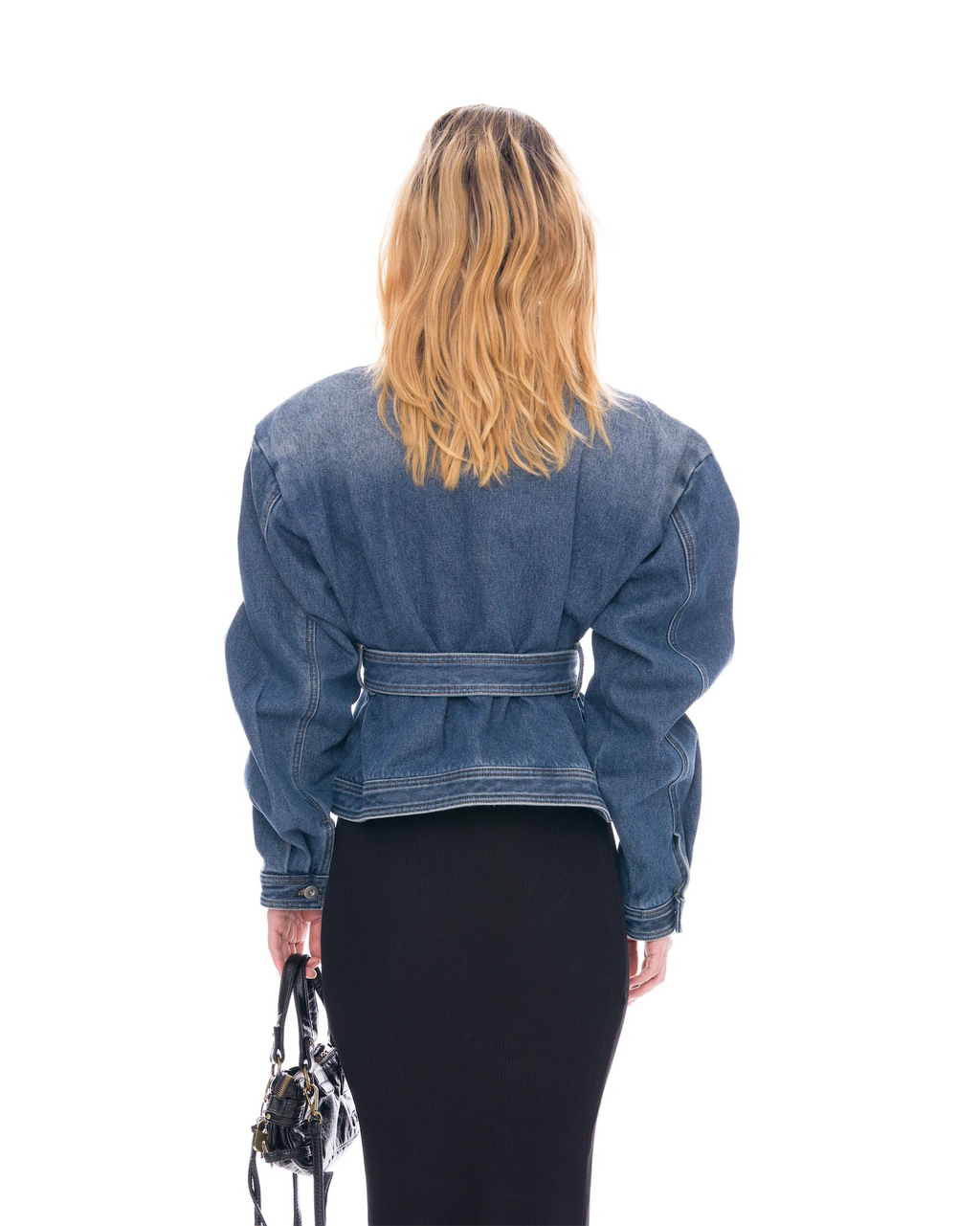 A woman shown from behind wearing a cropped blue denim jacket with a belted waist, paired with a black skirt, holding a small black handbag against a plain white background.