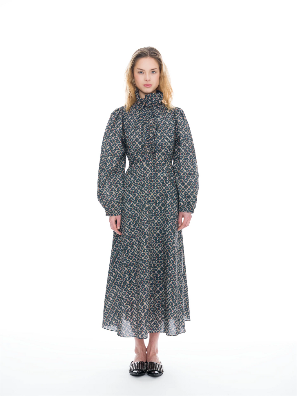 A model stands against a white studio background wearing a long-sleeved midi dress with a high mock neck, gathered waist, and an all-over geometric pattern in blue, orange, and gray.