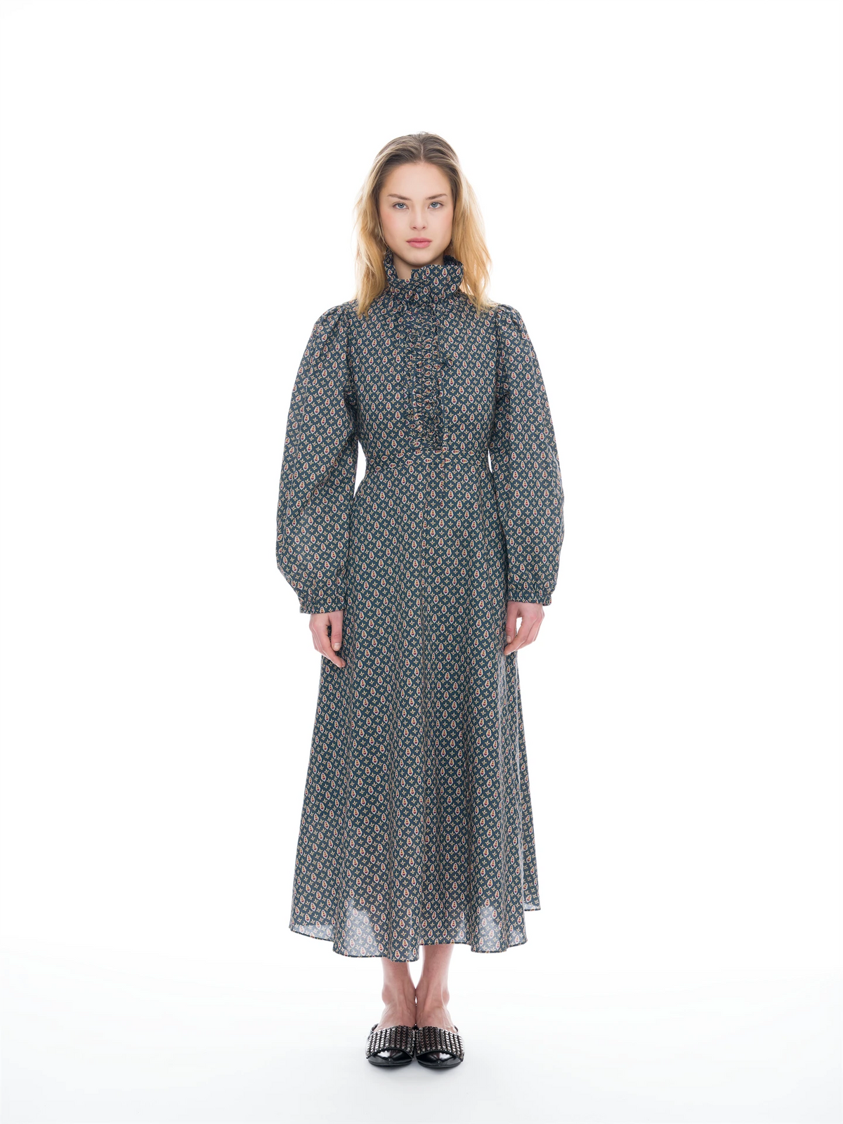 A model stands against a white studio background wearing a long-sleeved midi dress with a high mock neck, gathered waist, and an all-over geometric pattern in blue, orange, and gray.