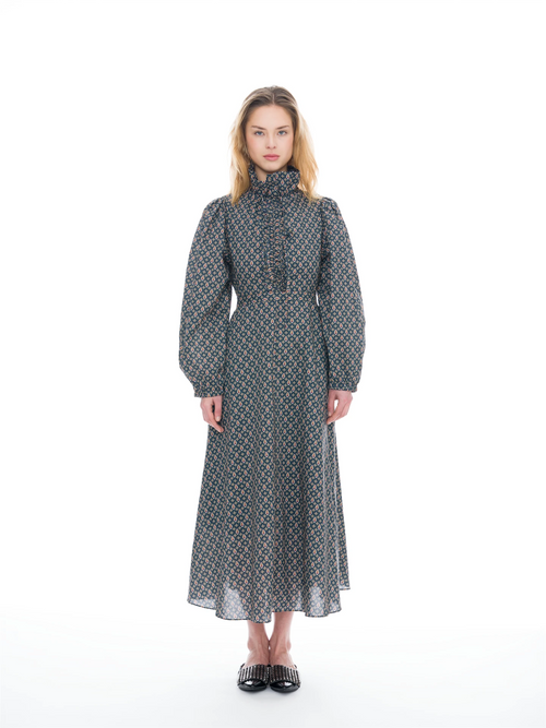 A model stands against a white studio background wearing a long-sleeved midi dress with a high mock neck, gathered waist, and an all-over geometric pattern in blue, orange, and gray.