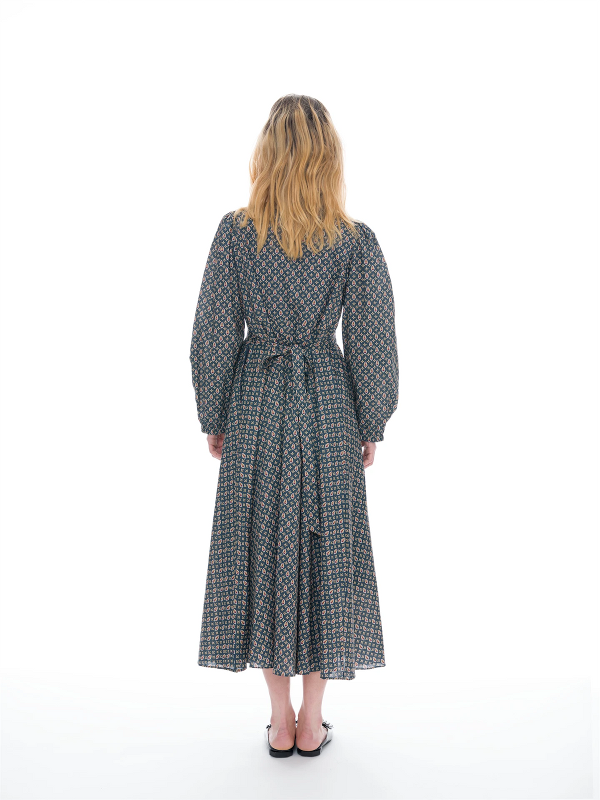 A woman viewed from behind wearing a long-sleeved midi dress with a geometric pattern in navy blue, beige, and orange, paired with black flat shoes, against a solid white studio background.