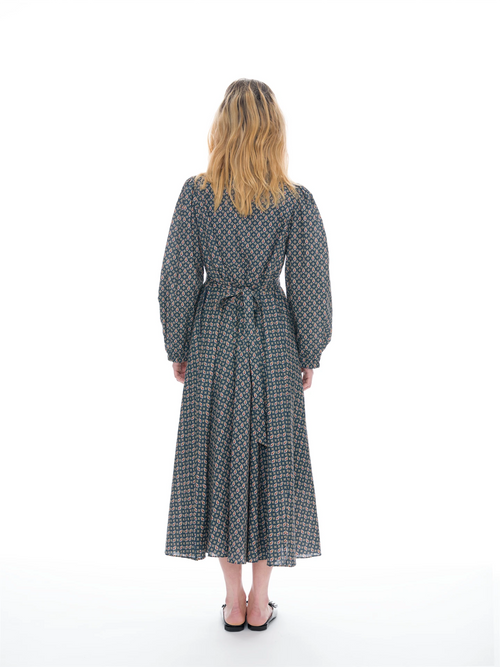A woman viewed from behind wearing a long-sleeved midi dress with a geometric pattern in navy blue, beige, and orange, paired with black flat shoes, against a solid white studio background.