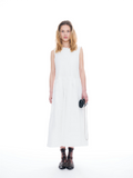 A woman modeling a sleeveless white midi dress with a high round neckline, paired with black strappy sandals and holding a small black camera bag, set against a seamless white studio background.