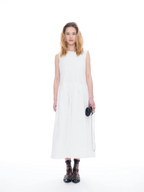 A woman modeling a sleeveless white midi dress with a high round neckline, paired with black strappy sandals and holding a small black camera bag, set against a seamless white studio background.