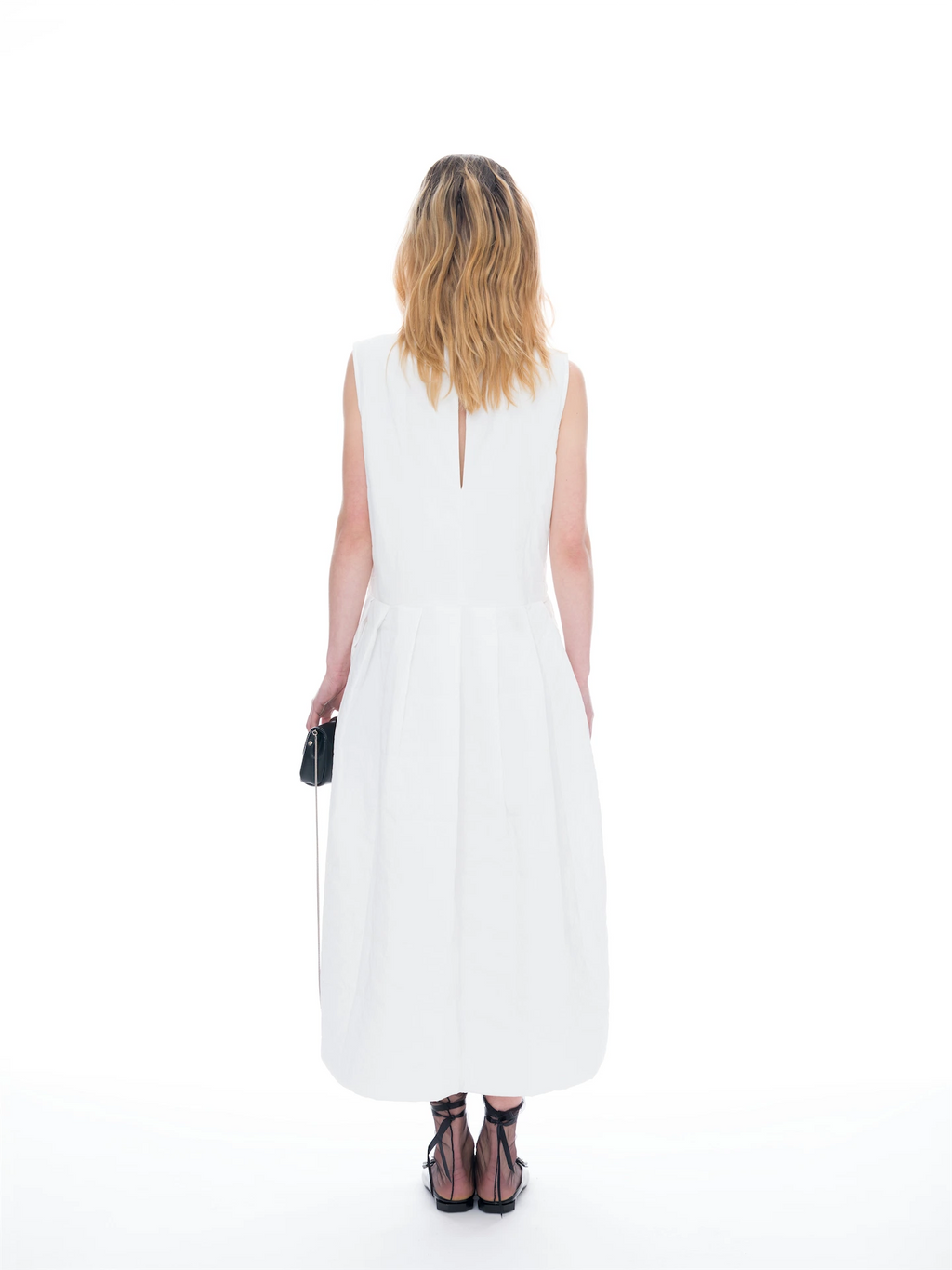 A woman viewed from behind wearing a sleeveless white midi dress with a small keyhole back cutout, paired with black strappy heels and holding a small black clutch on a seamless white background.