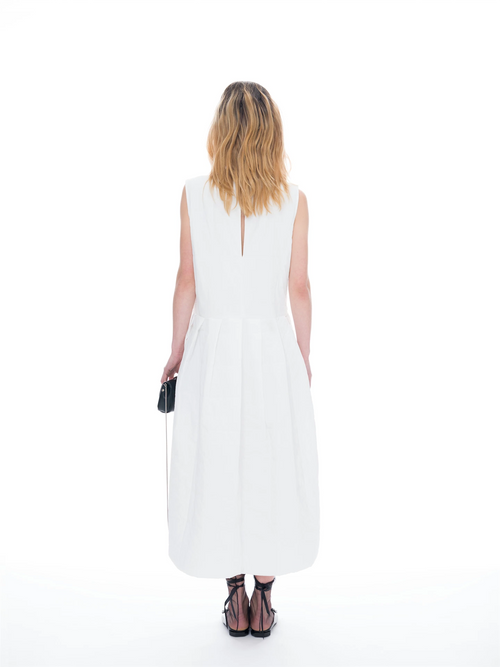 A woman viewed from behind wearing a sleeveless white midi dress with a small keyhole back cutout, paired with black strappy heels and holding a small black clutch on a seamless white background.