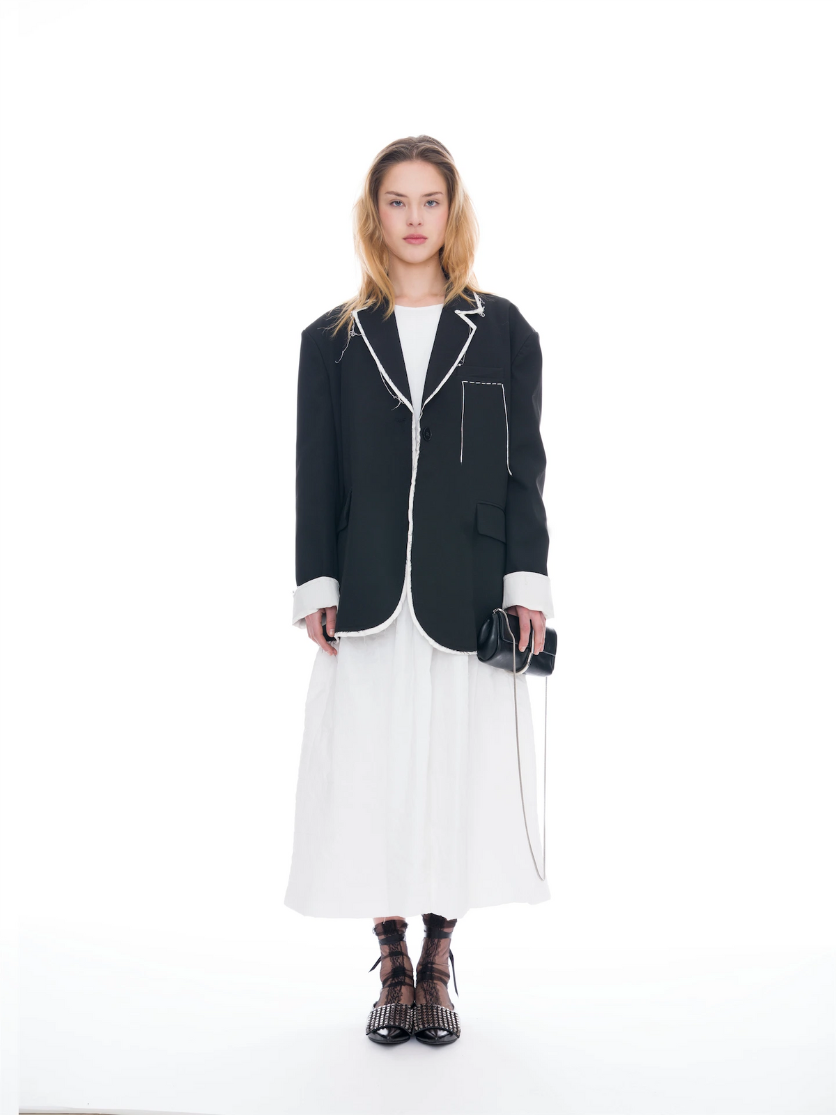 A model stands against a seamless white studio background wearing a black blazer with white contrast piping and cuffs, a white midi dress, black strappy high-heeled sandals, and holding a small black handbag.