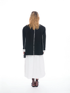 A woman viewed from behind wearing a black jacket with white piping and a white midi skirt, paired with black lace-up heels, against a seamless white studio background.