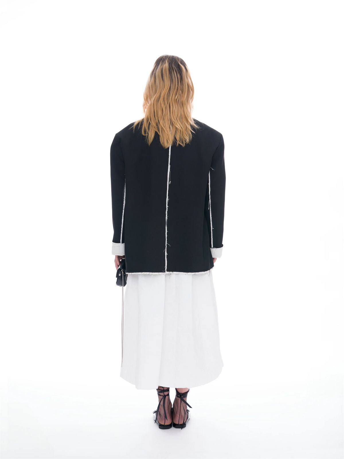 A woman viewed from behind wearing a black jacket with white piping and a white midi skirt, paired with black lace-up heels, against a seamless white studio background.