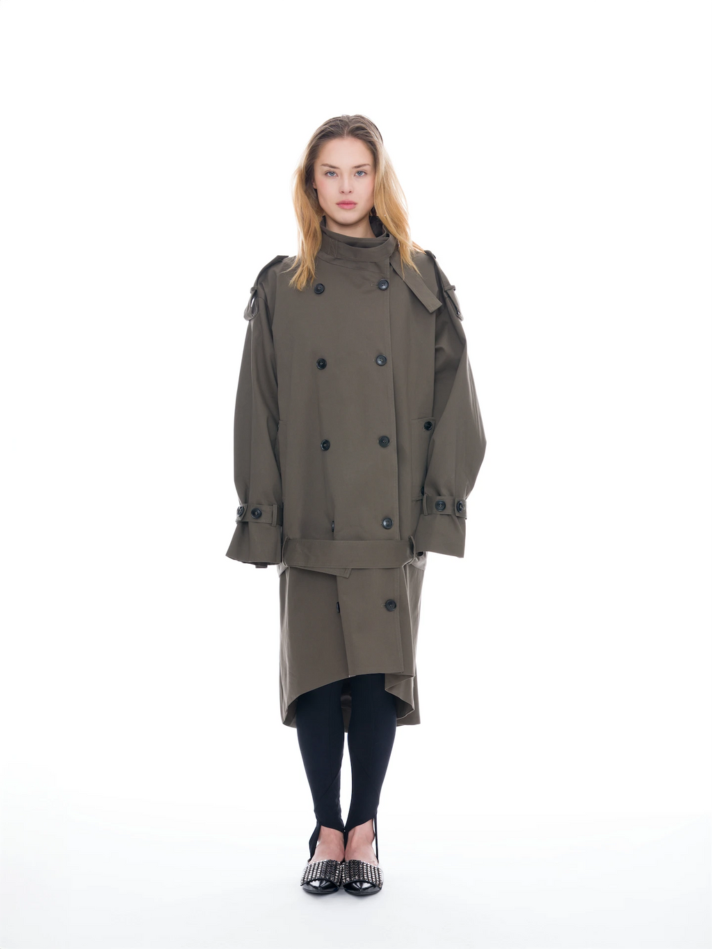 A model stands against a solid white background wearing a double-breasted olive green trench coat, black tights, and black shoes with silver studs.