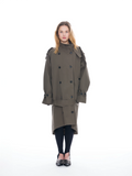 A model stands against a solid white background wearing a double-breasted olive green trench coat, black tights, and black shoes with silver studs.