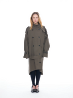 A model stands against a solid white background wearing a double-breasted olive green trench coat, black tights, and black shoes with silver studs.