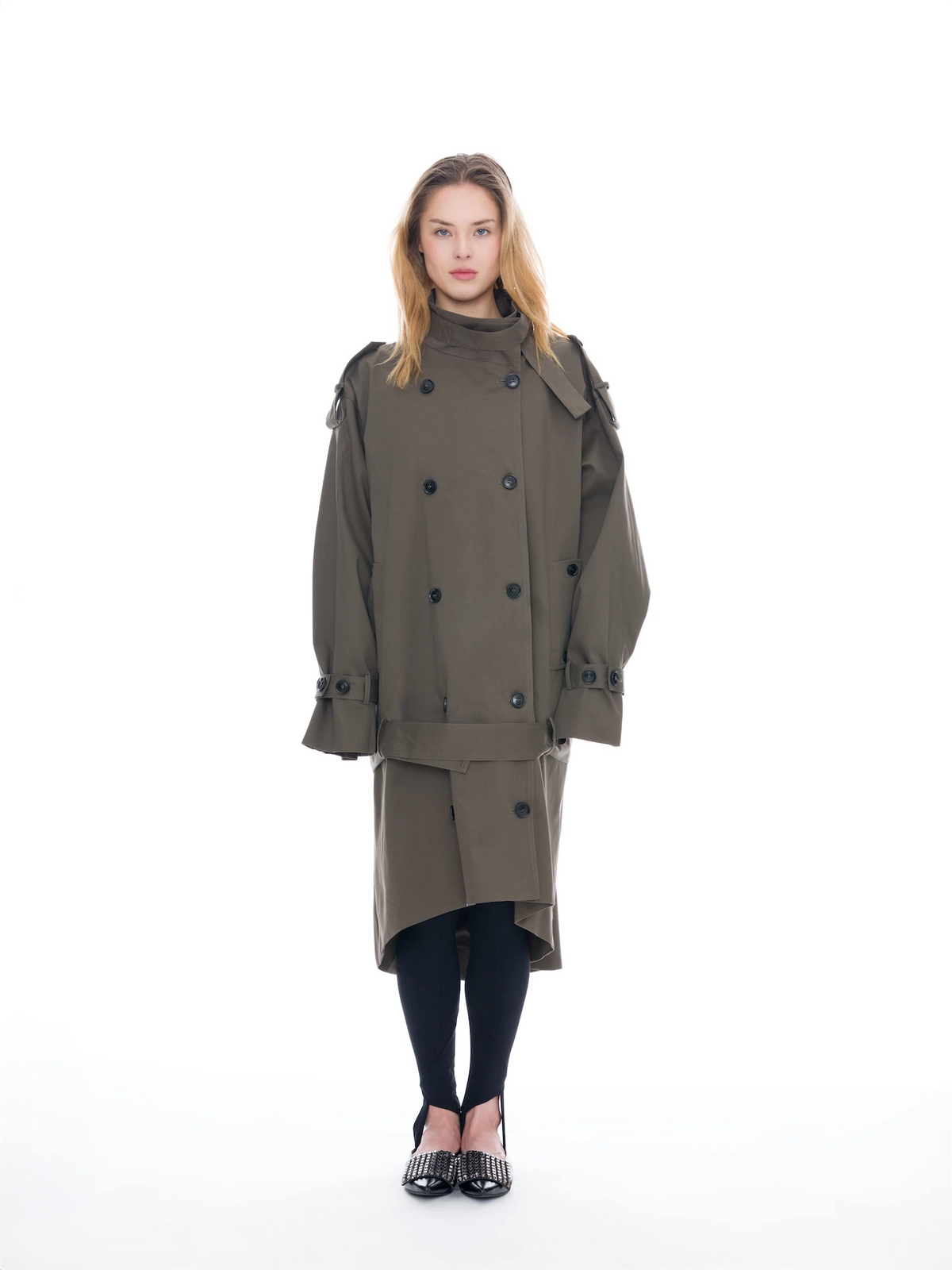 A model stands against a solid white background wearing a double-breasted olive green trench coat, black tights, and black shoes with silver studs.