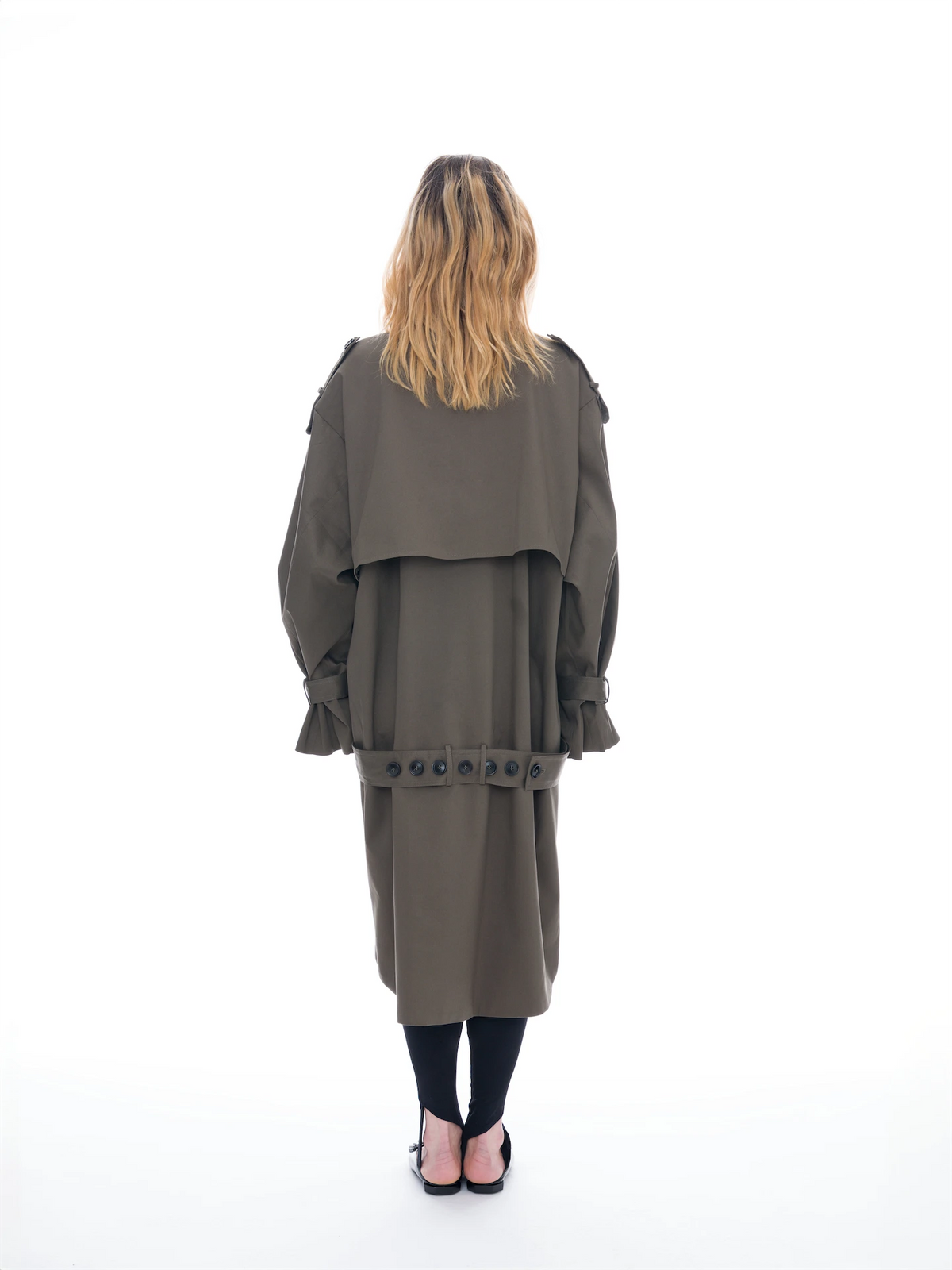 A model stands against a solid white background wearing a double-breasted olive green trench coat, black tights, and black shoes with silver studs.
