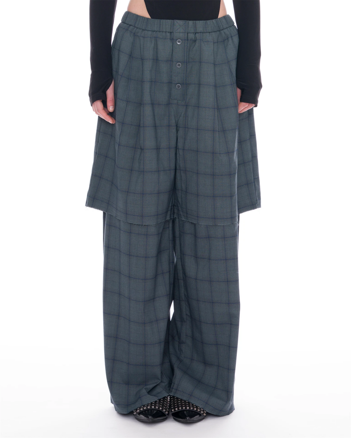 A model is shown from the waist down wearing wide-leg, high-waisted plaid trousers in a muted slate gray with a subtle blue undertone. The pants feature a button-up front and a draped, layered design.