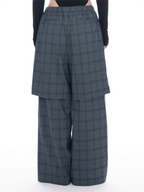 A model is shown from the waist down wearing wide-leg, high-waisted plaid trousers in a muted slate gray with a subtle blue undertone. The pants feature a button-up front and a draped, layered design.