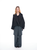 A model stands against a solid white studio background wearing a black oversized double-breasted jacket over a black top, paired with dark gray plaid wide-leg trousers.