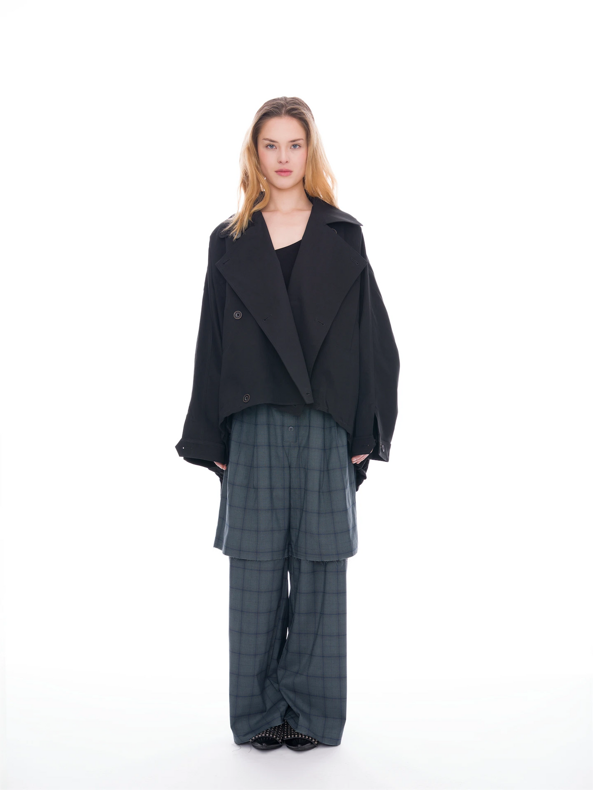 A model stands against a solid white studio background wearing a black oversized double-breasted jacket over a black top, paired with dark gray plaid wide-leg trousers.
