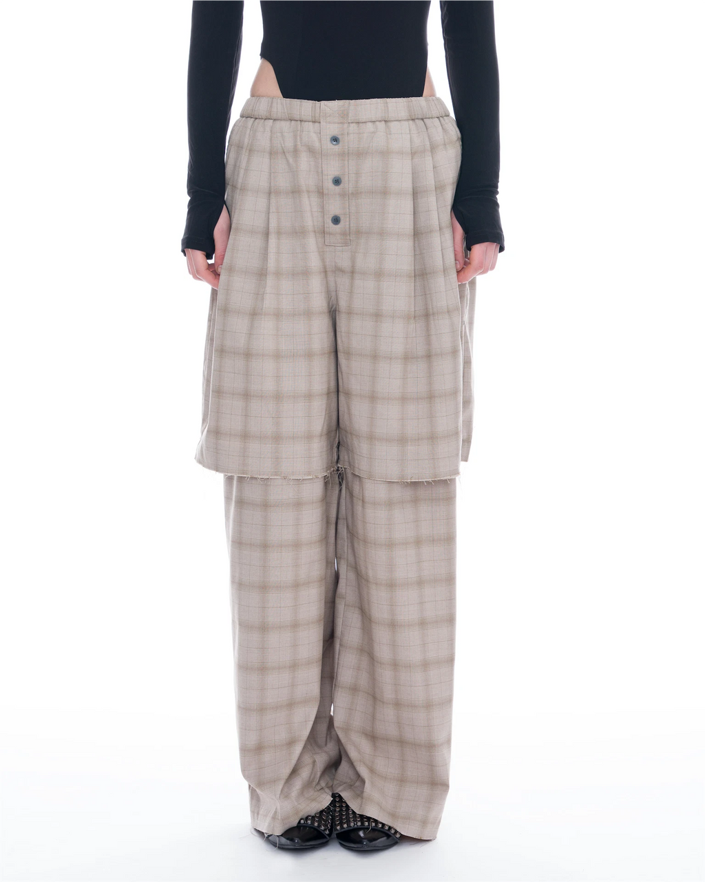 A model is shown from the waist down wearing beige and brown plaid wide-leg trousers with a button fly and a layered, draped front design. The model is also wearing a black long-sleeve top and black shoes.
