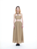 A model stands against a seamless white studio background wearing a two-piece outfit consisting of a beige tube top with white trim and a matching long beige skirt. She is also wearing a chunky brown cross necklace and black shoes.