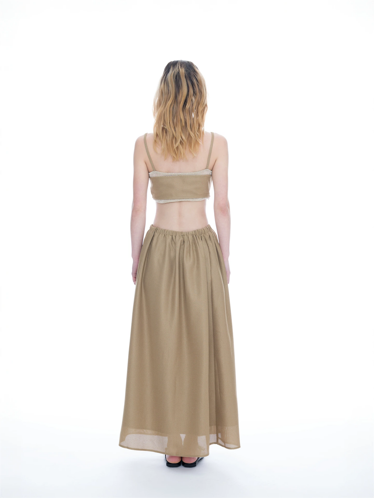 A woman viewed from behind wearing a beige two-piece set consisting of a cropped camisole top with thin straps and a matching high-waisted maxi skirt, set against a plain white studio background.