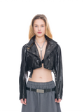 A model wearing a glossy black cropped moto jacket with silver snap details, styled with a black bralette, a silver pendant necklace, and a gray skirt with a black belt.