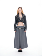 A model stands against a white studio background wearing a black cropped moto jacket, a dark gray wide-leg skirt, and a black belt with a silver buckle.