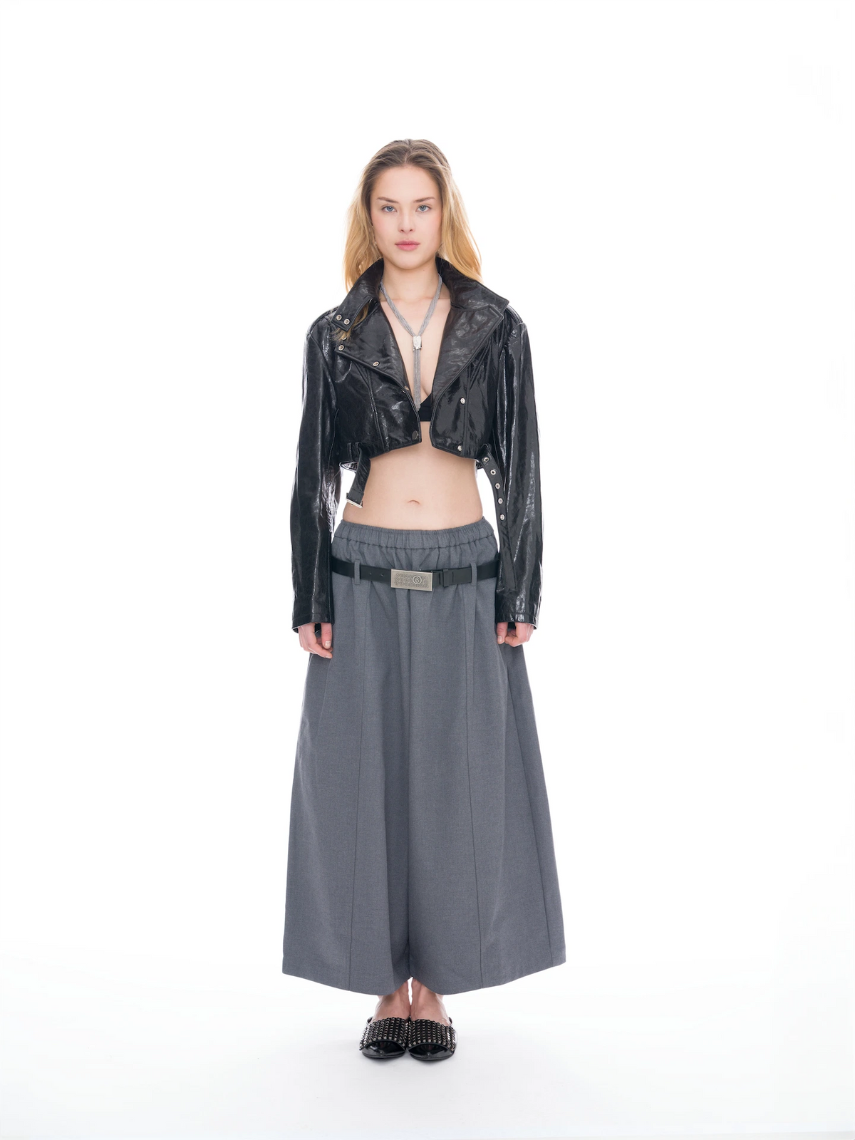 A model stands against a white studio background wearing a black cropped moto jacket, a dark gray wide-leg skirt, and a black belt with a silver buckle.