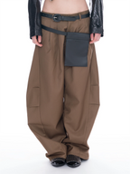 A model is shown from the waist down wearing wide-leg brown cargo pants with a black belt, a black utility pouch, and black studded shoes against a white background.