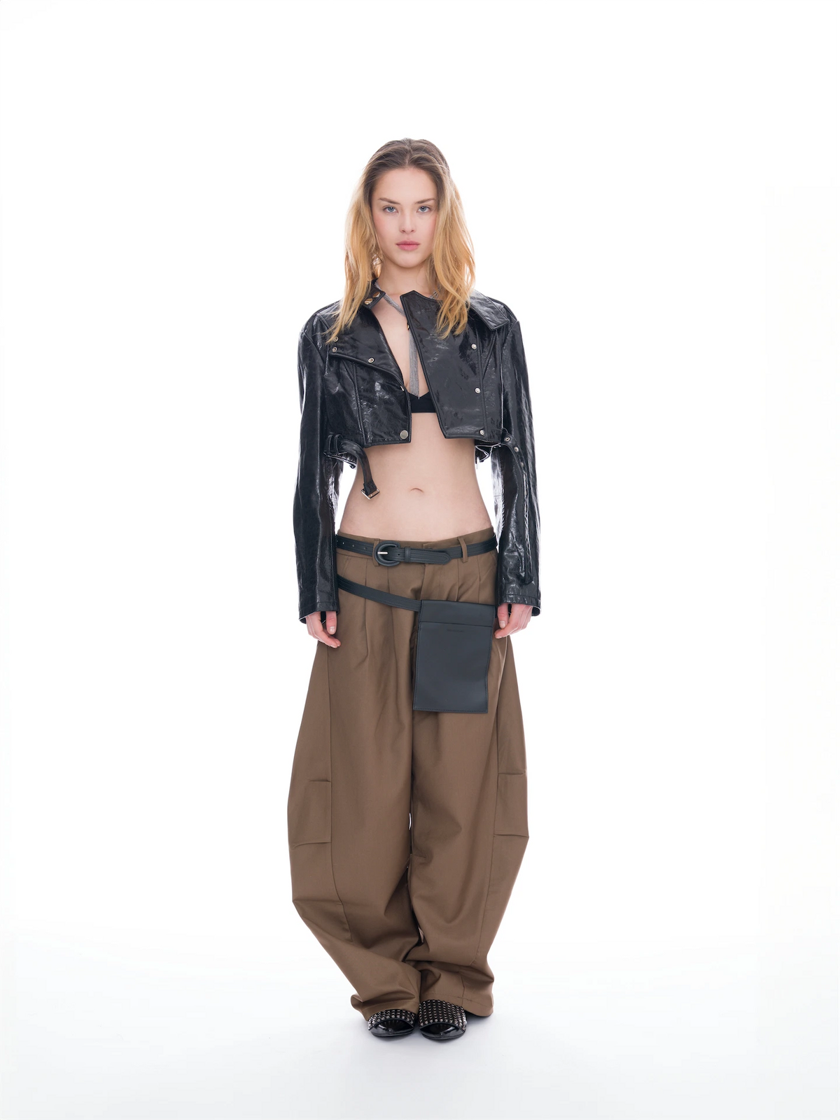 A model stands against a white studio background wearing a glossy black cropped jacket with a tie detail, loose brown cargo pants with a matching belt and pouch, and black shoes.