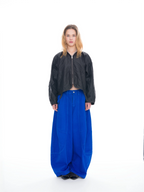 A model stands against a seamless white studio background wearing a black cropped zip-up jacket and extremely baggy royal blue wide-leg pants.