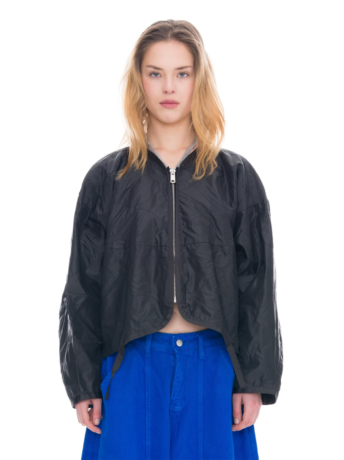 A model wearing a cropped black zip-up jacket with shiny textured panels and bright blue wide-leg pants, shot against a solid white background.