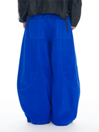 A person shown from the back wearing extremely baggy royal blue wide-leg pants with large patch pockets, paired with a black jacket, against a plain white background.