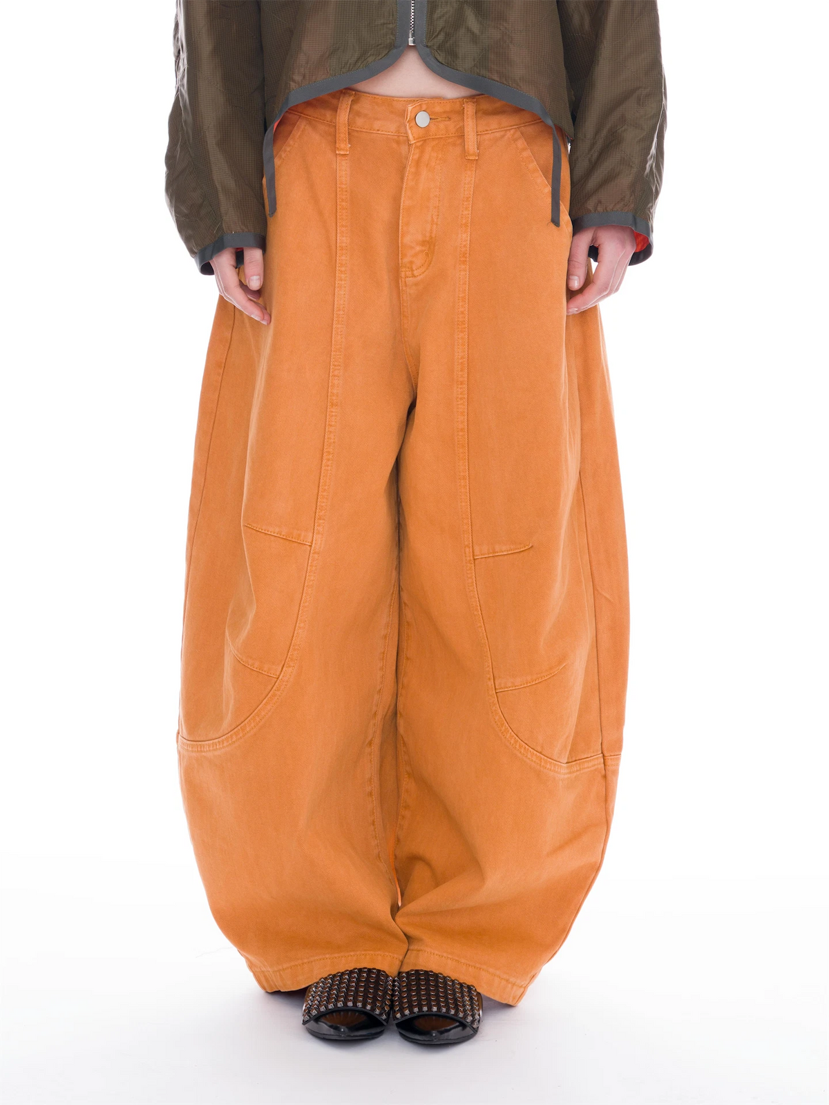 A person wearing oversized orange wide-leg cargo pants, an olive green cropped jacket, and black studded slide sandals, photographed against a plain white studio background.