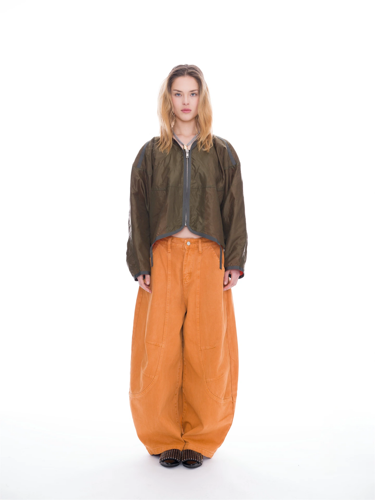 A model stands against a white studio background wearing a cropped olive green zip-up jacket, oversized orange wide-leg pants, and dark shoes.