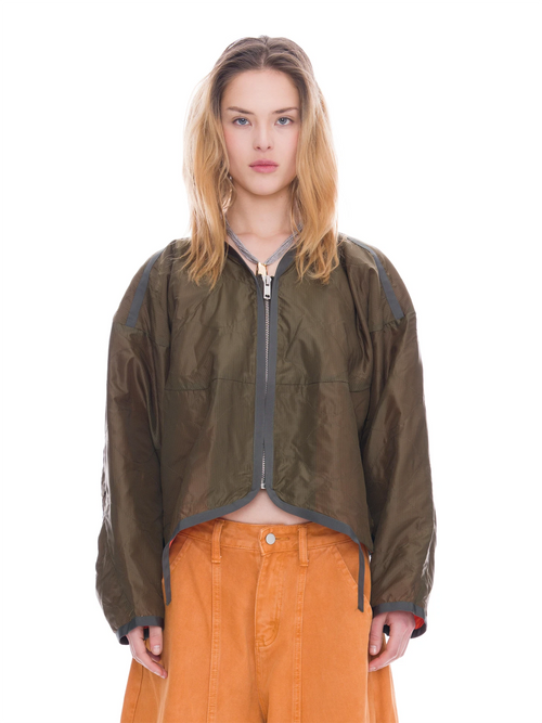 A model wearing an olive green cropped zip-up jacket with gray trim and orange pants, photographed against a solid white background.