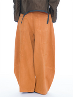 A person wearing oversized orange wide-leg cargo pants, an olive green cropped jacket, and black studded slide sandals, photographed against a plain white studio background.
