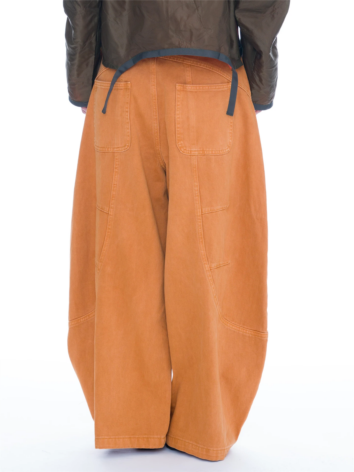 A person wearing oversized orange wide-leg cargo pants, an olive green cropped jacket, and black studded slide sandals, photographed against a plain white studio background.