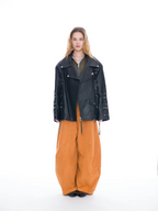 A model stands against a seamless white studio background wearing a black leather moto jacket with silver hardware, wide-leg orange-brown corduroy pants, and a dark olive top.