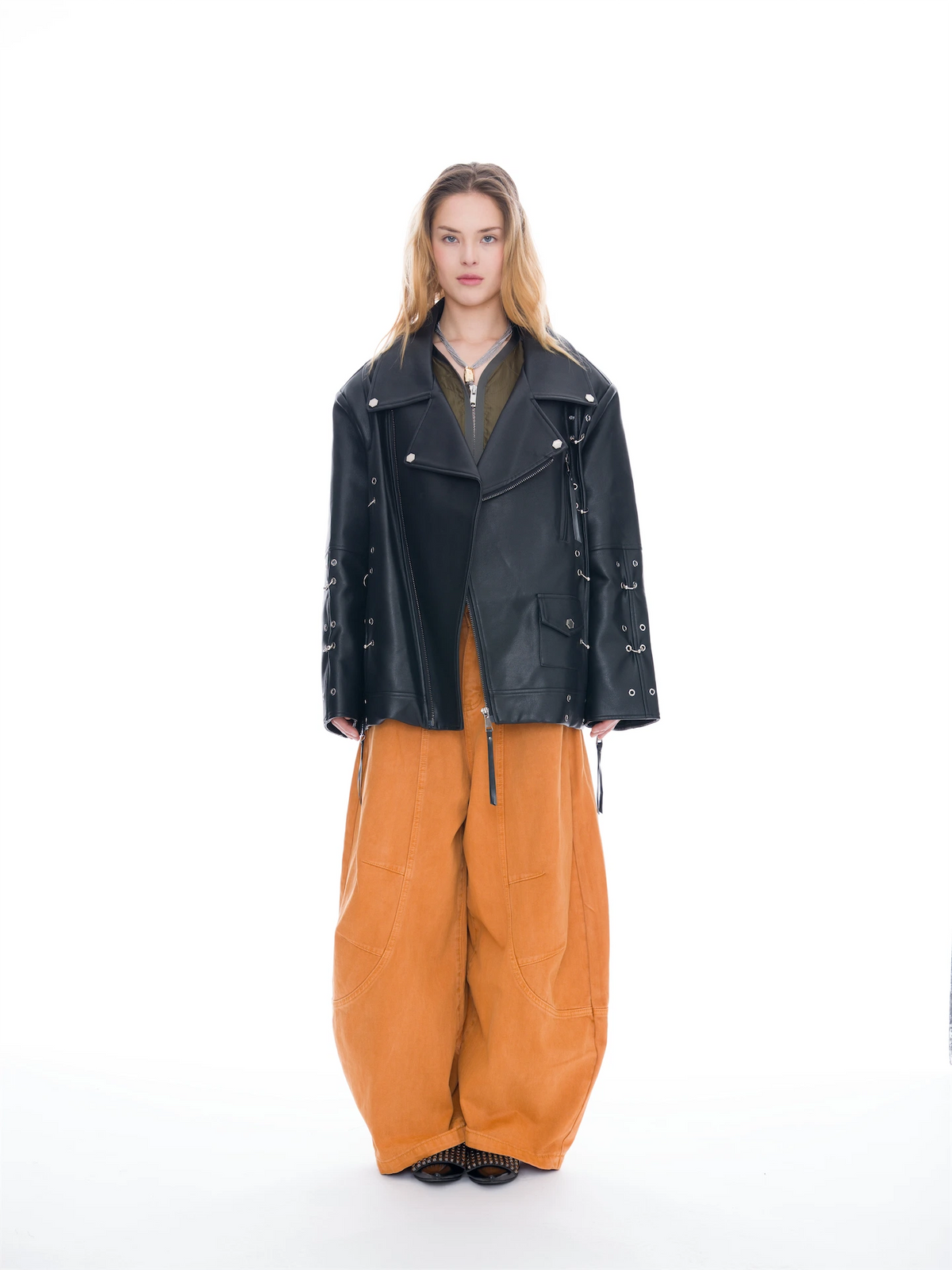 A model stands against a seamless white studio background wearing a black leather moto jacket with silver hardware, wide-leg orange-brown corduroy pants, and a dark olive top.