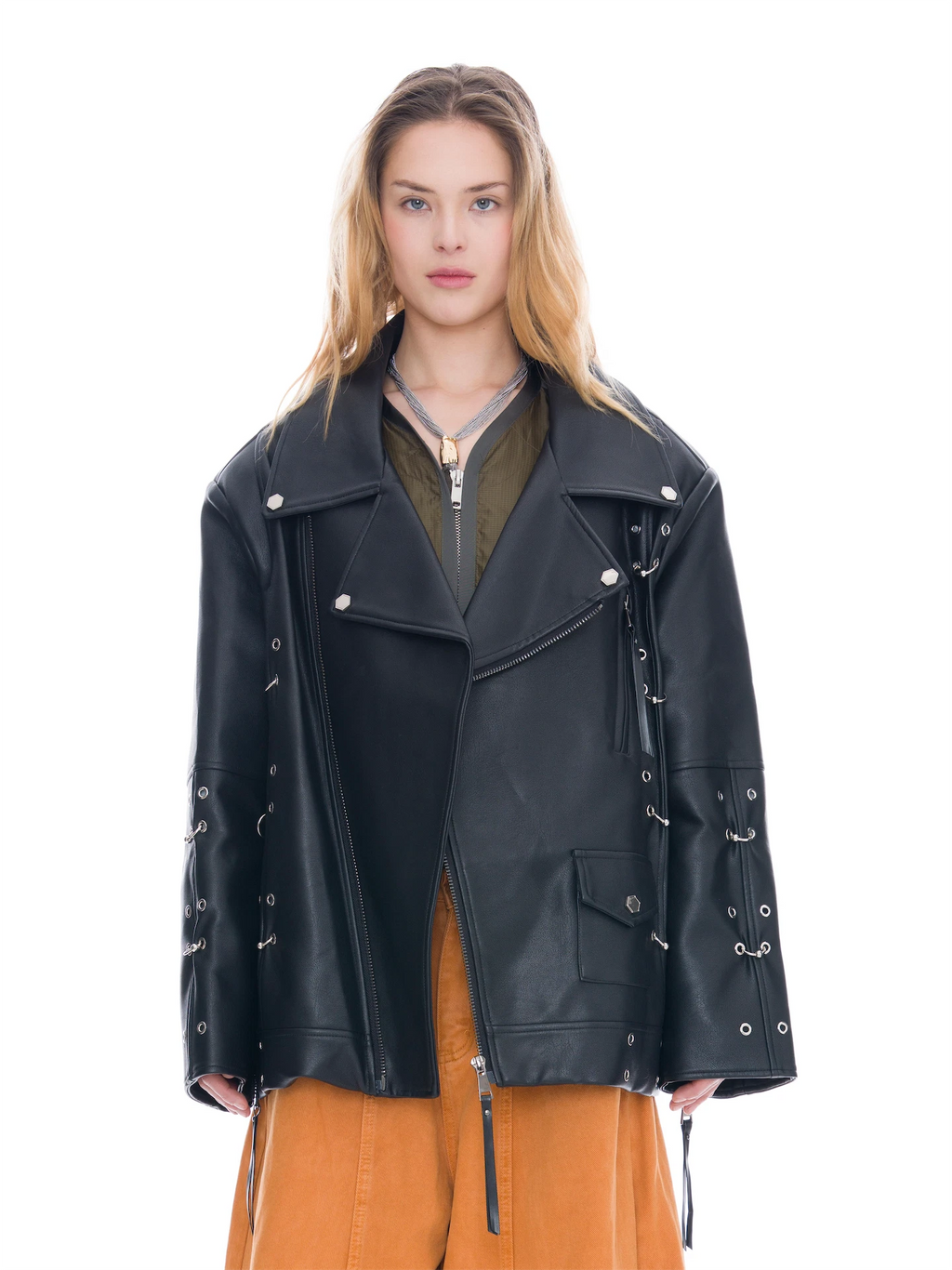 A model wearing a black faux-leather moto jacket with silver hardware, layered over an olive green zip-up top and orange-brown wide-leg pants, shot against a solid white background.