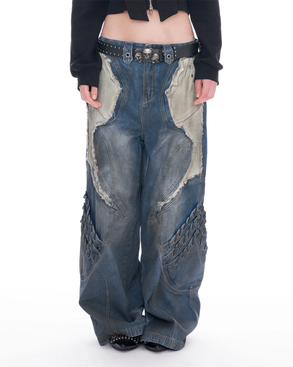 A person wearing extremely baggy blue denim pants with large distressed panels, chain detailing, and a studded belt, paired with a black cropped top against a white studio background.