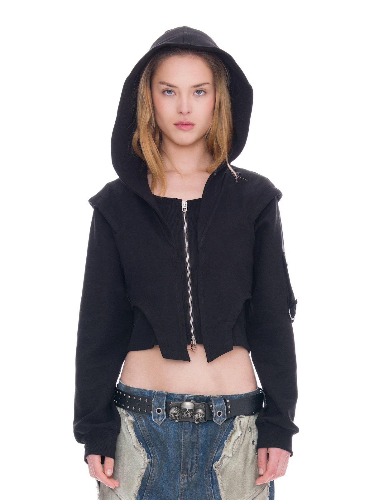 A model wearing a black cropped zip-up hoodie with a high hood, paired with distressed blue jeans and a black studded belt, against a solid white background.