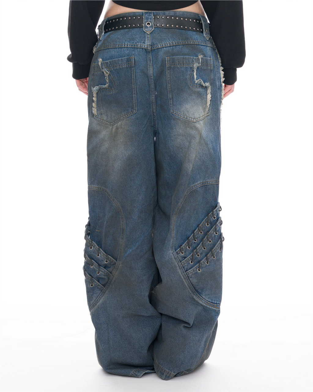 Rear view of a person wearing very baggy blue denim jeans with distressed stitching, decorative patch pockets, and metal eyelet embellishments on the knees, paired with a black belt and black long-sleeve top against a white studio background.