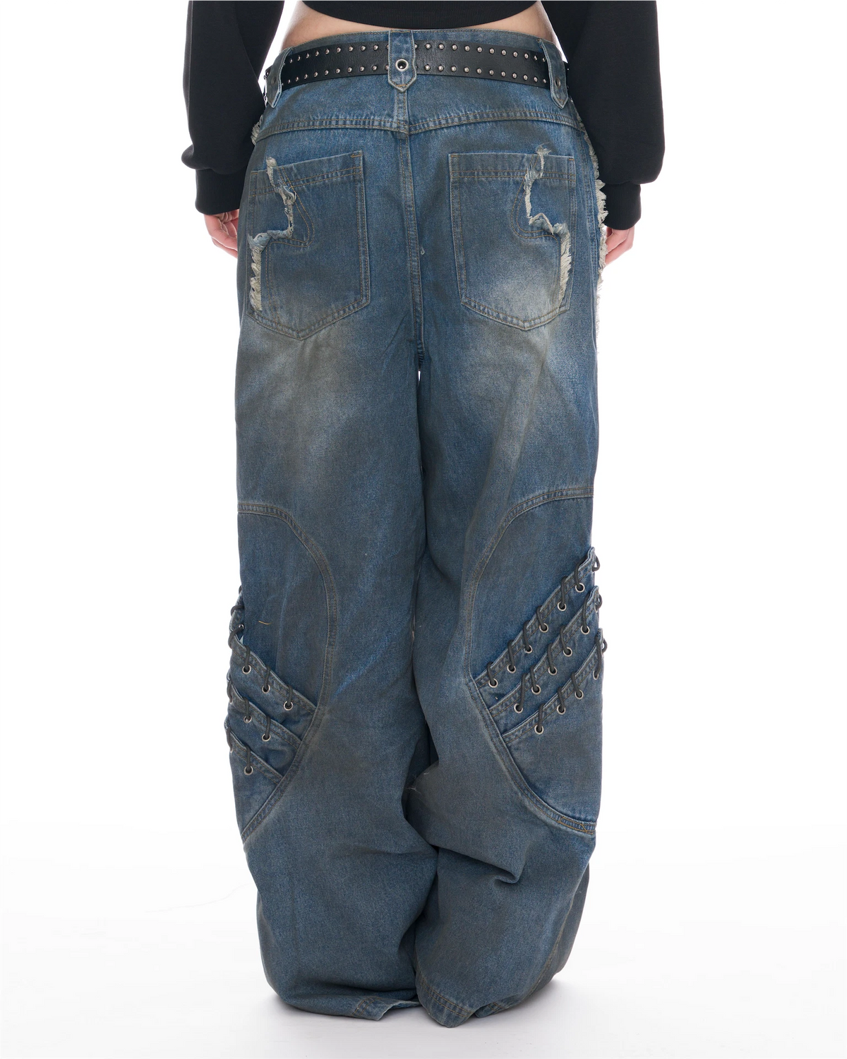 Rear view of a person wearing very baggy blue denim jeans with distressed stitching, decorative patch pockets, and metal eyelet embellishments on the knees, paired with a black belt and black long-sleeve top against a white studio background.