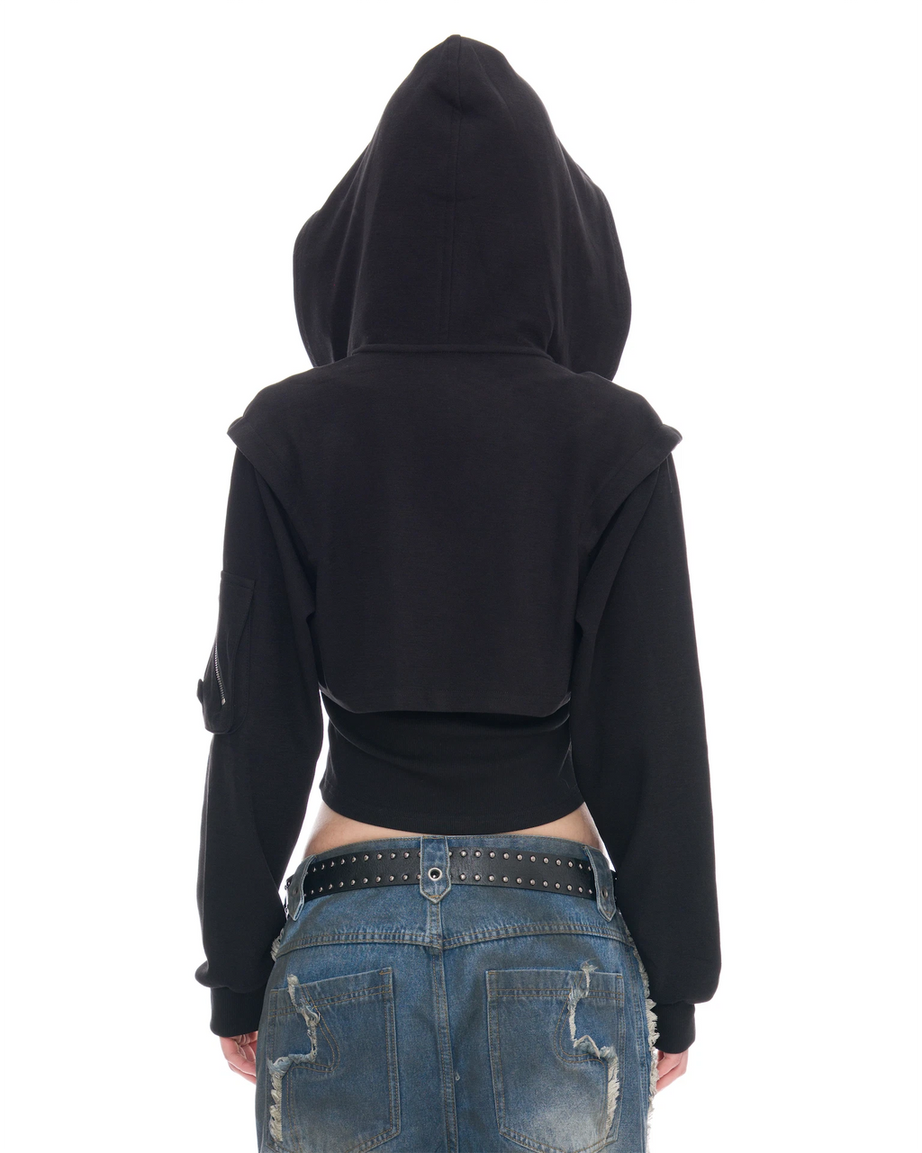 A person shown from the back wearing a black cropped hoodie with long sleeves and a high hood, paired with blue jeans and a black studded belt, against a plain white background.